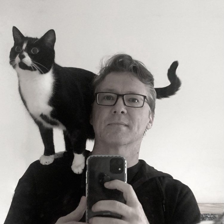The author taking a mirror selfie with a tuxedo cat on his shoulder looking intently at a moth it wants to eat.